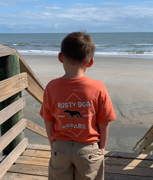 young boy wearing an orange Rusty Dog t-shirt. He is standing on a pier looking out over the ocean. 